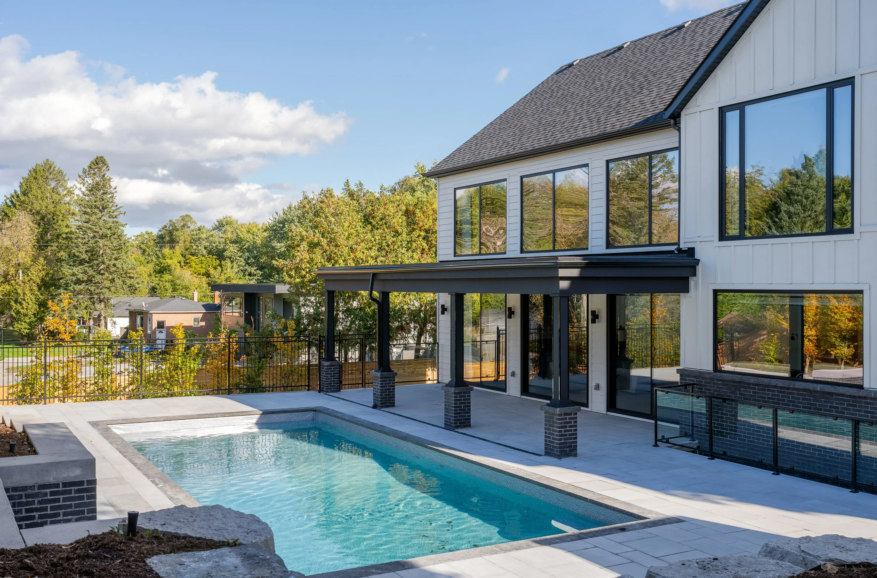 Rear of a white gabled residence with black window frames, opening onto a pool terrace in autumn.
