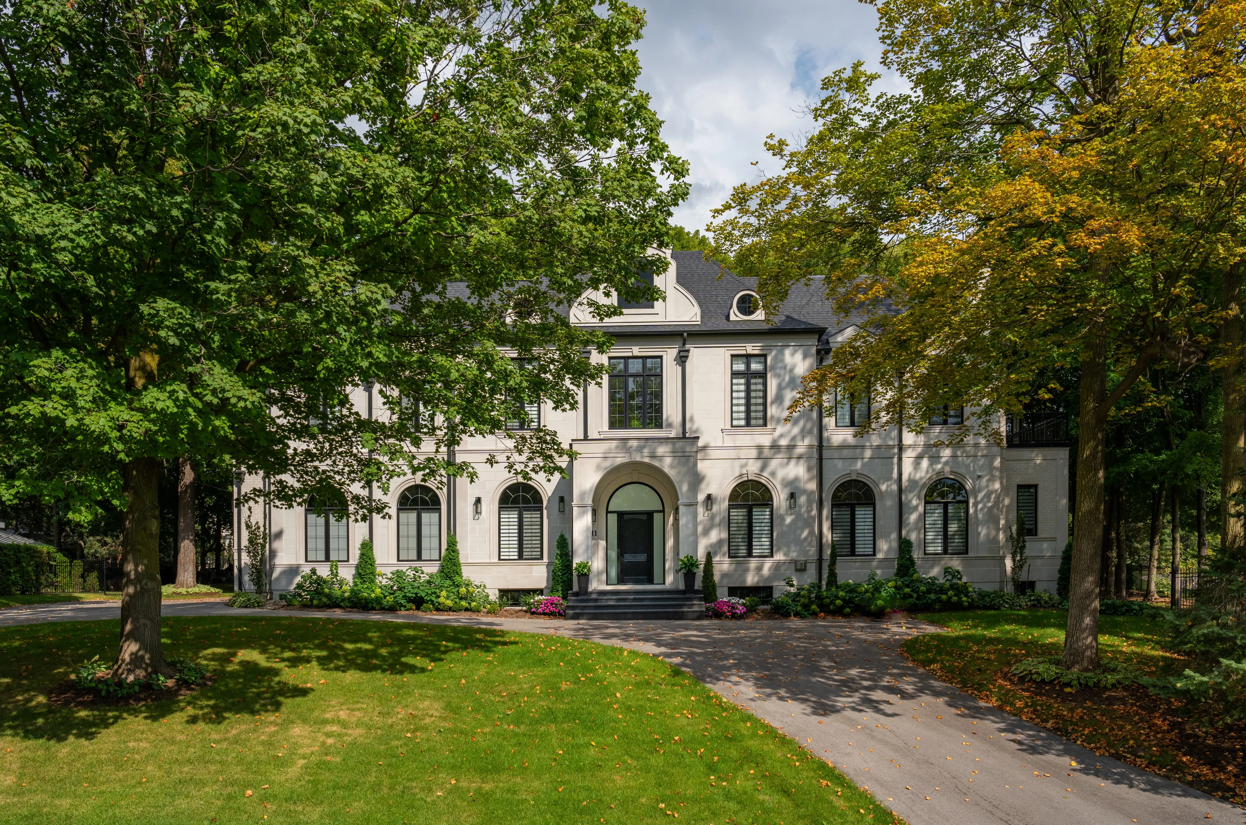 Limestone-clad residence with arched windows and a mansard roof, under mature trees.